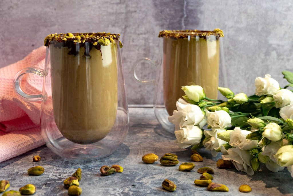 Pistachio Latte in a glass covered with chocolate and pistachio. Next to a pink tea towel, flowers and pistachios.